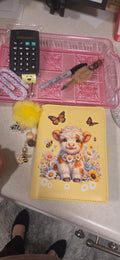 A yellow notebook or journal cover featuring a cute cartoon highland cow surrounded by flowers and butterflies, placed on a countertop next to a pink organizer tray, a calculator, and other small items.